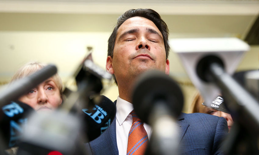 Photo of a middle-aged politician, eyes closed, chin raised, during a press conference bristling with microphones