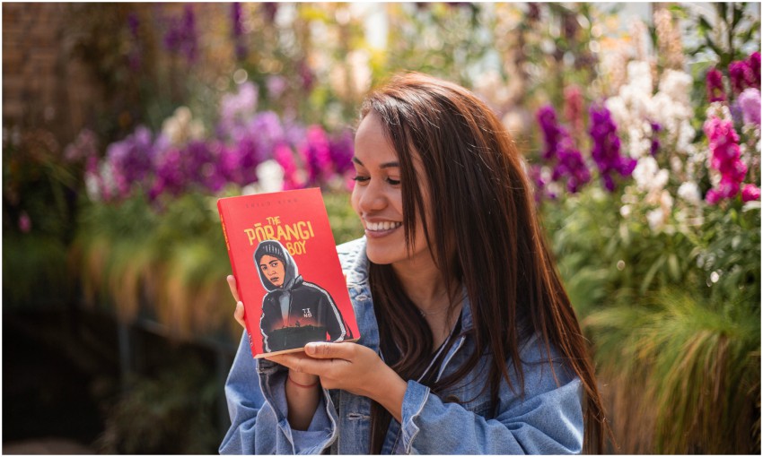 Young woman in garden holding novel The Pōrangi Boy, smiling