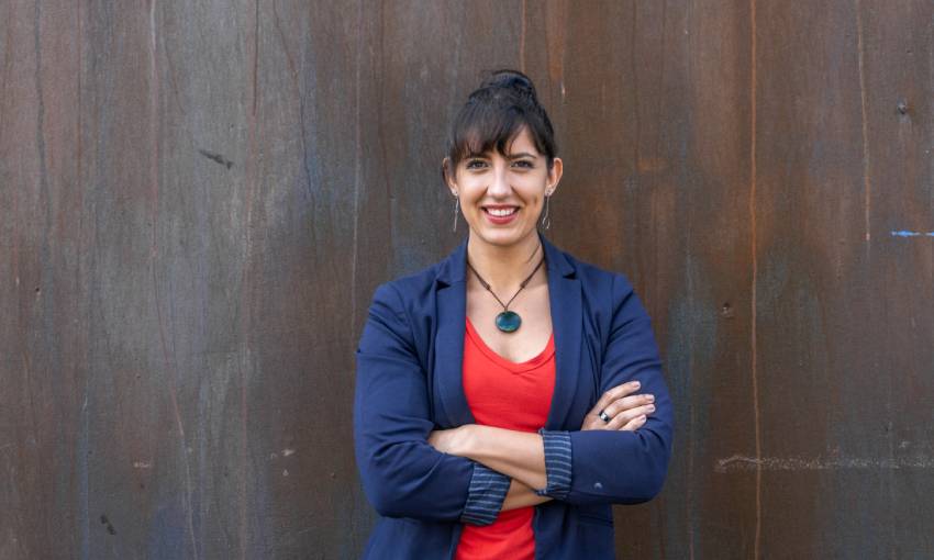 imche fourie wearing a blue blazer and red shirt, leaning against a wall with her arms crossed, smiling