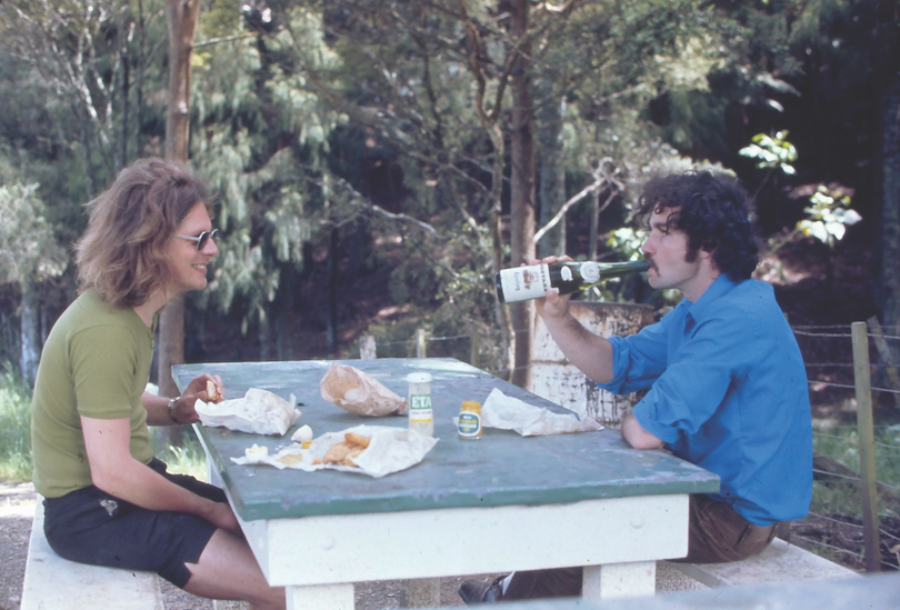 Two men at a picnic table, one swigging from a bottle, long hair and stubby shorts.