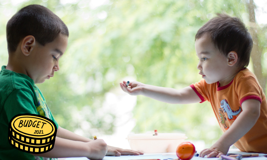 Two young mixed race boys coloring together at table next to window one year old is offering crayon to his older brother.