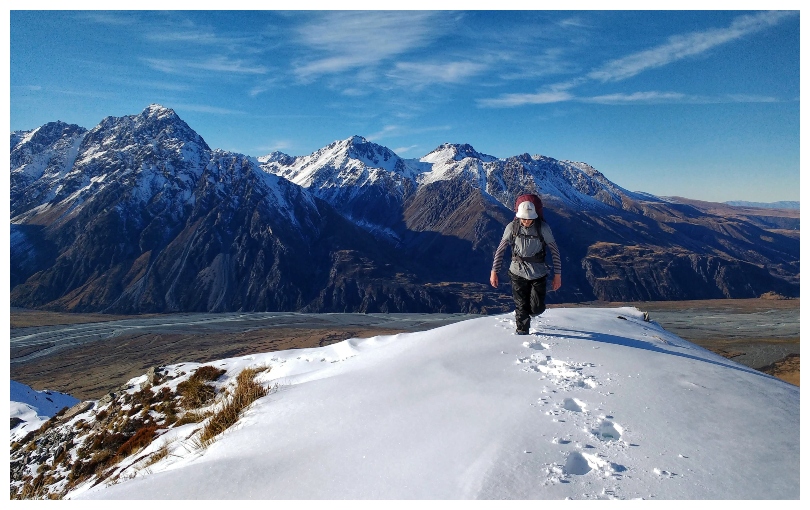 A beautiful clear day in the mountains, a young man trudging up a smooth stretch of snow towards camera.
