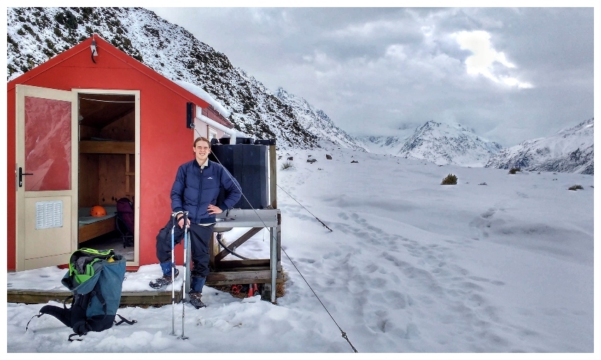 A young man in the snow, standing beside a bright red hut.