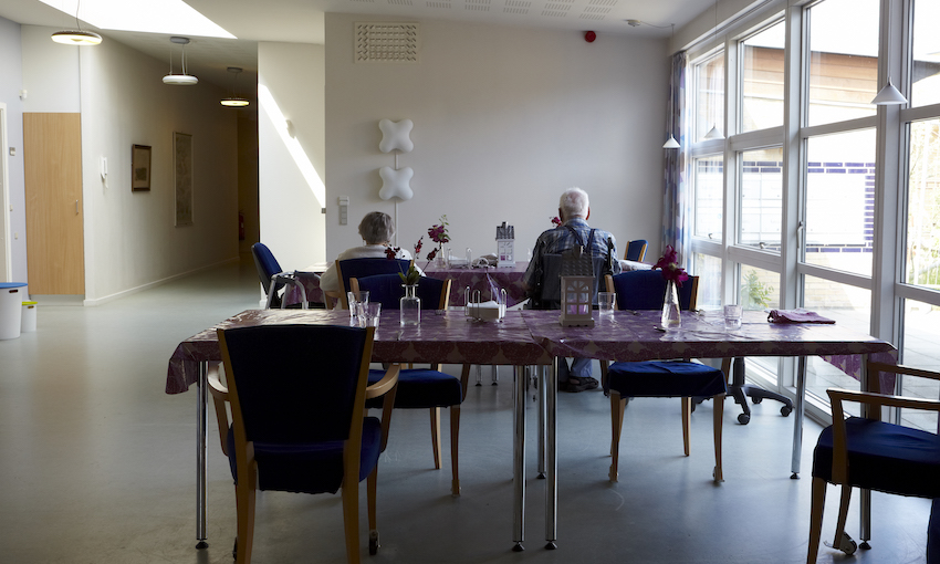 Interior of a rest home dining room; vinyl tablecloths, hard chairs, two residents seated.