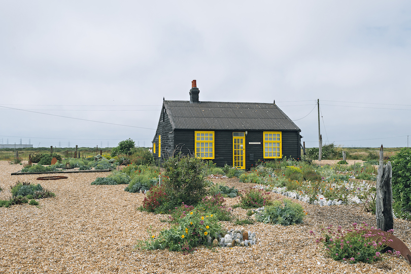 Striking black cottage with bright yellow trim. On a pebbled landscape, a garden of wildflowers.