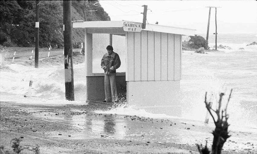 Old black and white image of a wild beach on Wellington's south coast, a man stands in a bus shelter beset by waves