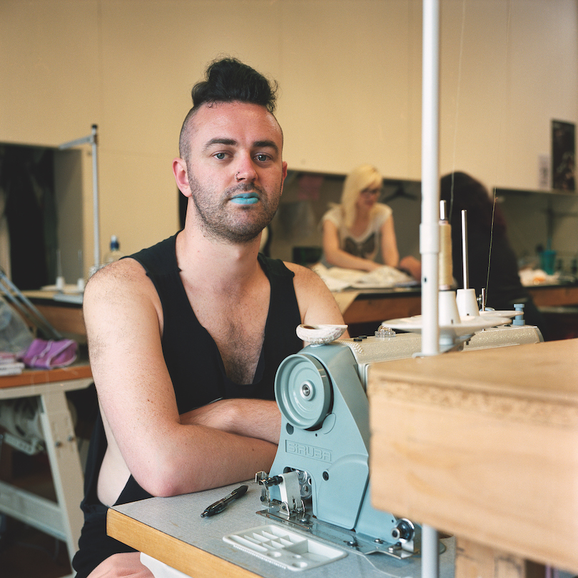 A young man wears a black singlet, blue lippy and a confident stare. He's seated at an industrial sewing machine, others (colleagues?) working in background.