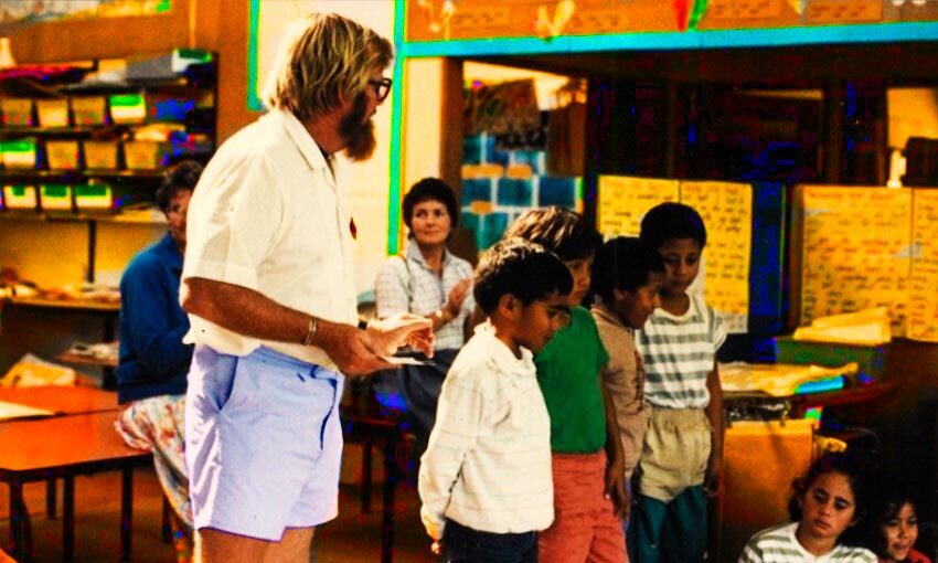 A 90s primary school classroom, male teacher with beard, short-sleeved shirt and walk shorts.