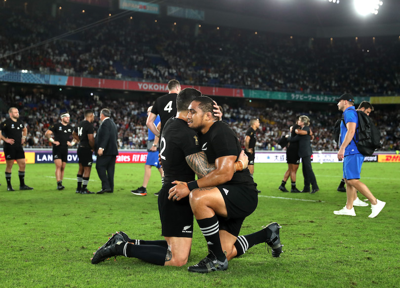 Sonny Bill Williams and Ofa Tu'ungafasi in the foreground, in All Blacks kit, embracing, on their knees; in the background, aftermath of a rugby match.