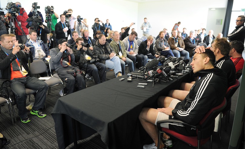 A sea of white male journalists, seated, face Sonny Bill Williams and a few other players (obscured) at a press conference. He looks uncomfortable, sitting awkwardly and scratching his head.